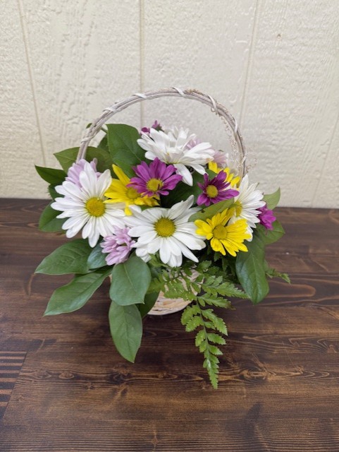 Basket of yellow, purple and white daisies and greenery on a wooden table.