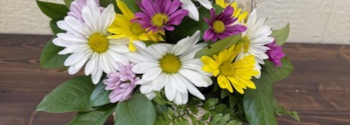 Basket of yellow, purple and white daisies and greenery on a wooden table.
